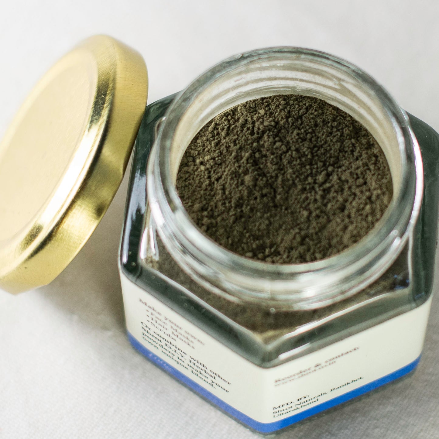 Hexagonal glass jar with gold lid containing Shrot Indigo powder on a white background. Top angle with lid off.
