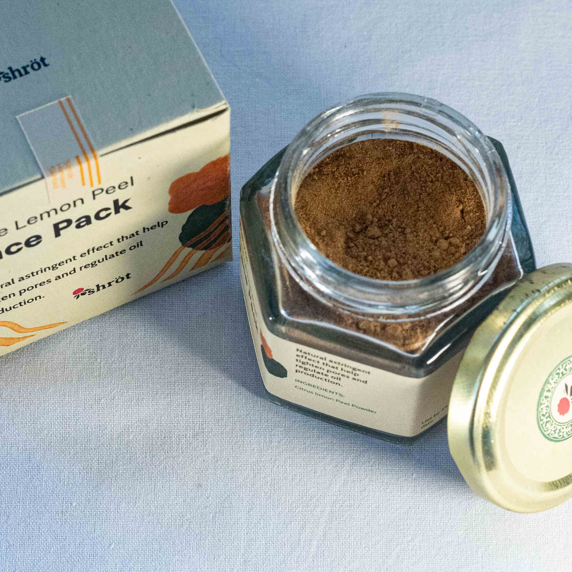 Jar of brown Lemon powder with a box labeled 'Lemon Peel Face Pack' by Shrot natural cosmetics on a light blue background